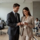 A professional office environment in modern Austria, two colleagues in business attire sharing a subtle, affectionate moment by the coffee machine in an open-plan workspace. Soft natural light filters through large windows, other employees blurred in the background focused on their tasks. The scene conveys warmth, discretion, and everyday romance without disrupting the professional atmosphere. Clean, editorial photographic style with realistic details and neutral color palette.