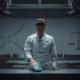 A somber, editorial-style courtroom illustration in muted blues and grays: a young male chef in kitchen uniform stands alone at the center, looking concerned. One of his hands is gently scanned by a glowing biometric hand vein reader, while two security cameras above him (labeled 10 and 23) point directly at his workspace in a professional kitchen. Faint broken chains and a balance scale appear subtly in the background, symbolizing power imbalance and privacy rights. The atmosphere is serious and reflective, with soft dramatic lighting, no text, logos, or branding. Cinematic composition, high detail, editorial photography style.